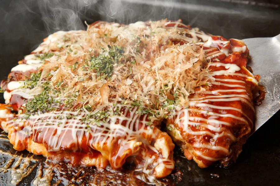 鉄板の上で焼かれた熱々のお好み焼きをヘラで持ち上げている画像